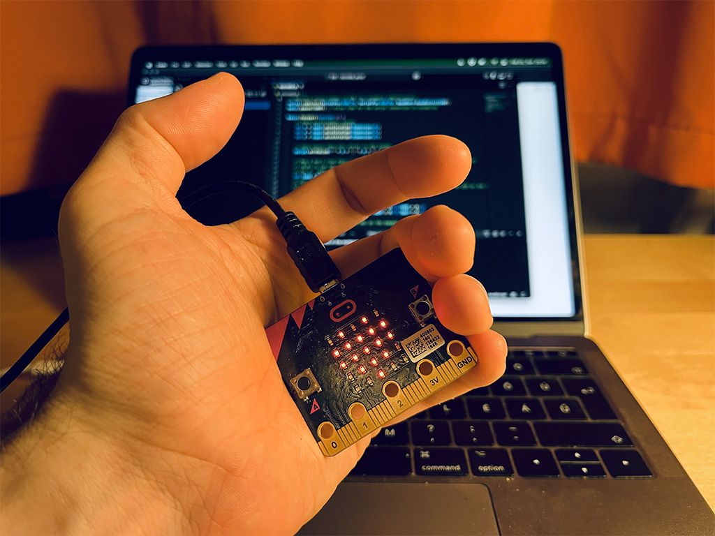 A micro:bit in my palm, plugged into a computer, with a heart displayed on its LEDs